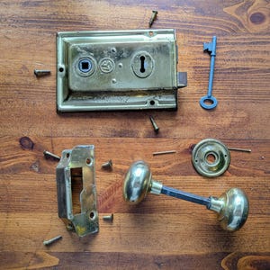 May include: An antique brass door lock set, disassembled on a wooden surface. The set includes a rectangular lock plate, a door knob, a strike plate, a key, and several screws. The components are arranged for restoration.