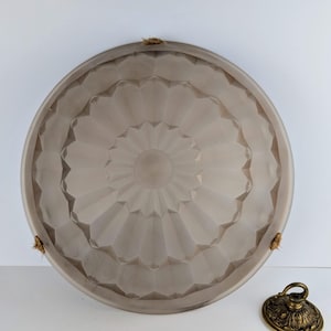 May include: A large, round, gray-toned glass ceiling light fixture with a decorative, geometric pattern. The fixture has three small rope attachments and sits next to a decorative brass ceiling cap.