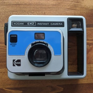 Puede incluir: Una cámara instantánea Kodak EK2 gris y negra con un paquete de película azul y blanco. La cámara tiene un flash incorporado y un visor.