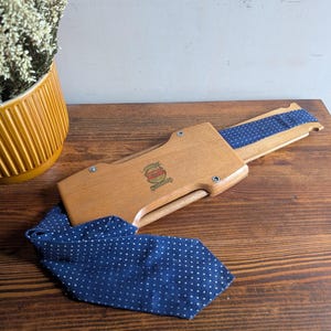 Peut inclure: Un étui à cravate en bois avec une cravate bleu marine à pois blancs. L'étui est ouvert, révélant une section de la cravate à l'intérieur. L'étui porte un logo sur le devant et est posé sur une surface en bois.