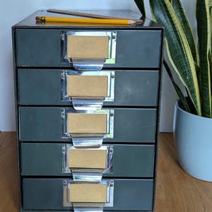 Puede incluir: Un archivador de metal vintage con seis cajones. Cada cajón tiene un portaetiquetas y una manija de metal. El archivador está pintado de color verde oscuro.