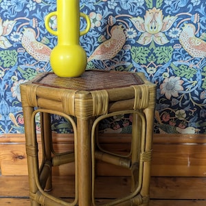 May include: A small, hexagonal, brown wicker side table with a yellow ceramic vase on top. The table has a woven top and sits on a wooden floor.