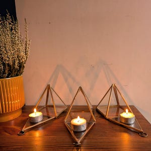 Puede incluir: Tres candelabros geométricos con velas encendidas sobre una superficie de madera. Los candelabros son de metal dorado y vidrio, con forma de pirámides. A la izquierda se ve una maceta de cerámica amarilla con una planta.