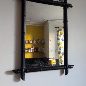 May include: A black bamboo framed mirror with a chain hanging from the top. The mirror reflects a room with a yellow wall and a shelf with teacups.
