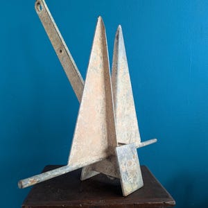 Puede incluir: Un ancla de metal desgastado con un diseño geométrico único. El ancla presenta dos grandes uñas triangulares y un vástago largo y angulado. El metal tiene un aspecto oxidado y envejecido, lo que sugiere que es una pieza antigua. Se encuentra sobre una superficie de madera oscura sobre un fondo azul.