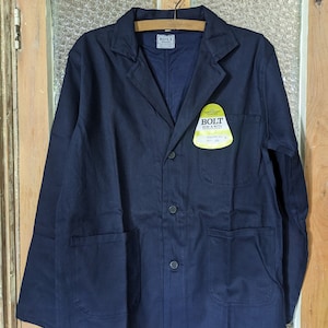 May include: A dark blue work jacket with a button closure and two large pockets. The jacket has a label that reads "Bolt Brand" with the text "Guaranteed Workwear" underneath.