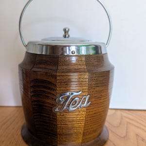 May include: A wooden tea caddy with a silver lid and handle. The caddy is shaped like a barrel and has the word "Tea" inscribed on the front.