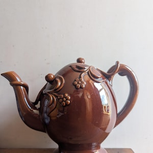 May include: A brown ceramic teapot with a floral design. The teapot has a rounded body and a curved spout. The handle is attached to the side of the teapot.