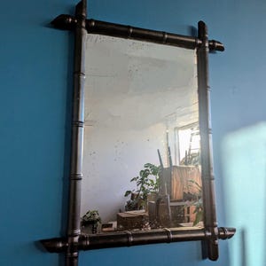 May include: A black bamboo framed mirror with a rectangular shape. The mirror is hanging on a blue wall and reflects a room with plants and furniture.