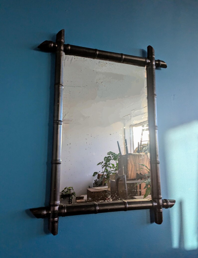 May include: A black bamboo framed mirror with a rectangular shape. The mirror is hanging on a blue wall and reflects a room with plants and furniture.