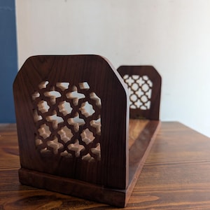 Puede incluir: Un sujetalibros de madera marrón oscuro con intrincados patrones geométricos calados. El sujetalibros tiene una base rectangular y dos soportes verticales, cada uno con aberturas decorativas. La veta de la madera es visible.