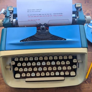 Puede incluir: Una máquina de escribir vintage azul y crema con papel blanco en el carro. El texto en el papel dice: "Máquina de escribir Imperial Safari, Fabricada en 1971 en Portugal". También se ven una taza y un platillo azules y un lápiz amarillo.