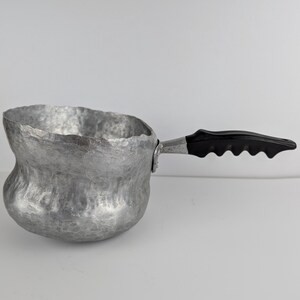 May include: A small, hammered aluminium saucepan with a black plastic handle. The pan has a unique, textured surface and a curved shape. The handle is riveted to the side of the pan.