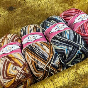 Può includere: Quattro gomitoli di filato in diverse combinazioni di colori. Ogni gomitolo è etichettato "alize Superwash 100g" e presenta una varietà di colori, tra cui marrone, nero, bianco, blu, rosso e grigio.