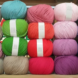May include: A collection of yarn balls in various colours, including yellow, orange, brown, pink, green, red, purple, blue, grey, and white. The yarn balls are arranged in rows on a wooden surface.