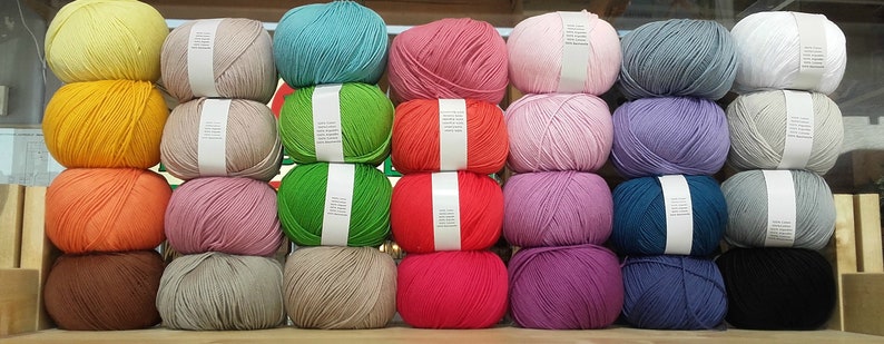 May include: A collection of yarn balls in various colours, including yellow, orange, brown, pink, green, red, purple, blue, grey, and white. The yarn balls are arranged in rows on a wooden surface.
