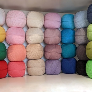 Puede incluir: Una colección de 24 ovillos de lana en varios colores, incluyendo blanco, amarillo, verde, rojo, rosa, beige, marrón, azul y morado. Los ovillos de lana están dispuestos en filas en un estante blanco.