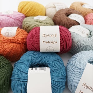 Op de afbeelding: Een verzameling kleurrijke garenbollen, waaronder oranje, rood, blauw, groen en geel. Het garen is gelabeld met "Rosários 4 Madragoa Fibras Naturais Natural".