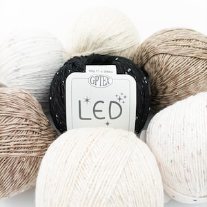 Op de afbeelding: Vijf bollen garen in verschillende kleuren, waaronder zwart, wit, beige en bruin. De zwarte bol garen heeft een label met de tekst "GPTEX LED 50g e = 200m".