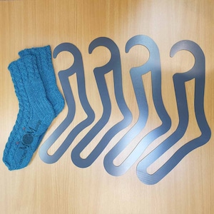 May include: Five black sock-shaped hangers for storing knitted socks. The hangers are made of a sturdy material and have a smooth finish. A pair of teal blue knitted socks with a cable pattern are also shown in the image.