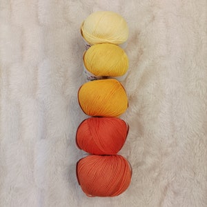 May include: Five balls of yarn stacked on top of each other. The top ball is a pale yellow, the next is a bright yellow, the next is a bright orange, and the bottom two are a burnt orange colour.