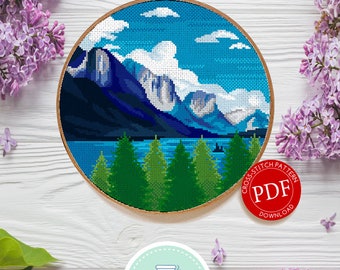 May include: A cross stitch pattern featuring a mountain landscape with a lake and trees. The pattern is in a round hoop and the text "Cross Stitch Pattern PDF Download" is on a red circle.