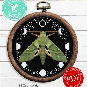 May include: A cross stitch pattern featuring a green moth with white accents, surrounded by white moons and stars on a black background. The pattern is framed in a brown wooden hoop and labeled "14 Count Aida" and "Cross Stitch Pattern PDF Download".