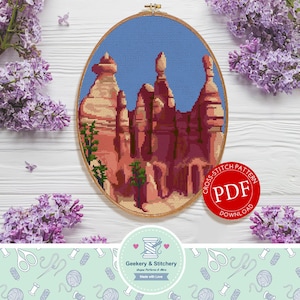 May include: Cross stitch pattern PDF download for a colorful landscape with red rock formations and a blue sky. The pattern is in a hoop and surrounded by purple flowers.