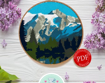 May include: A cross-stitch pattern PDF download for a mountain landscape with a lake in the foreground. The pattern is displayed on a wooden surface with flowers and a pair of scissors.