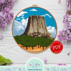 May include: A cross-stitch pattern of Devils Tower National Monument in Wyoming, a large rock formation with a blue sky and white clouds in the background. The pattern is in a round wooden embroidery hoop. The image also includes a red circle with the text "Cross Stitch Pattern PDF Download".