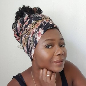 Op de afbeelding: Een vrouw met donker krullend haar draagt een zwarte en roze bloemenhoofddoek met gouden bladeren. Ze draagt een zwart tanktop en heeft een gouden ketting.