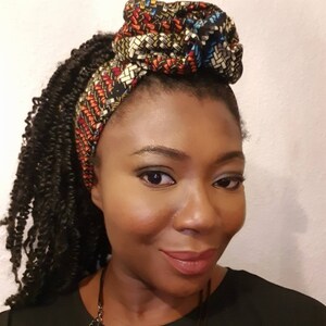 Pu&ograve; includere: Una donna con i capelli castano scuro raccolti in trecce indossa una fascia per capelli con motivo nero e marrone con un grande nodo in cima. Indossa una camicia nera e una collana d'argento.