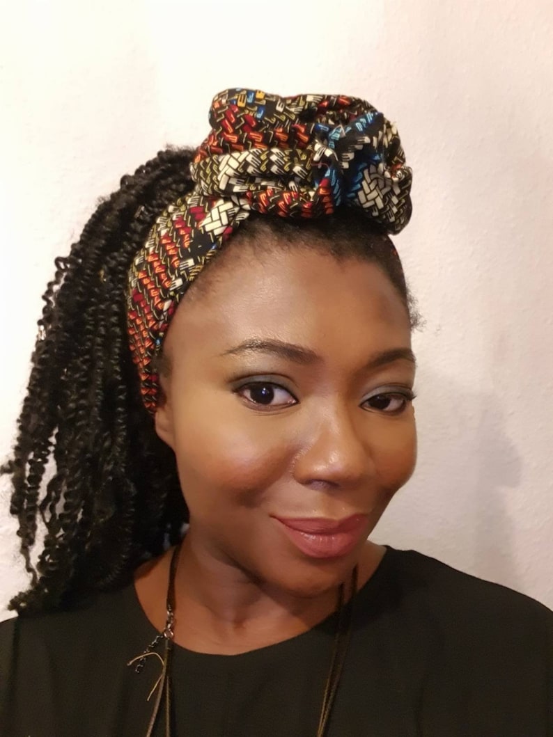 Pu&ograve; includere: Una donna con i capelli castano scuro raccolti in trecce indossa una fascia per capelli con motivo nero e marrone con un grande nodo in cima. Indossa una camicia nera e una collana d'argento.