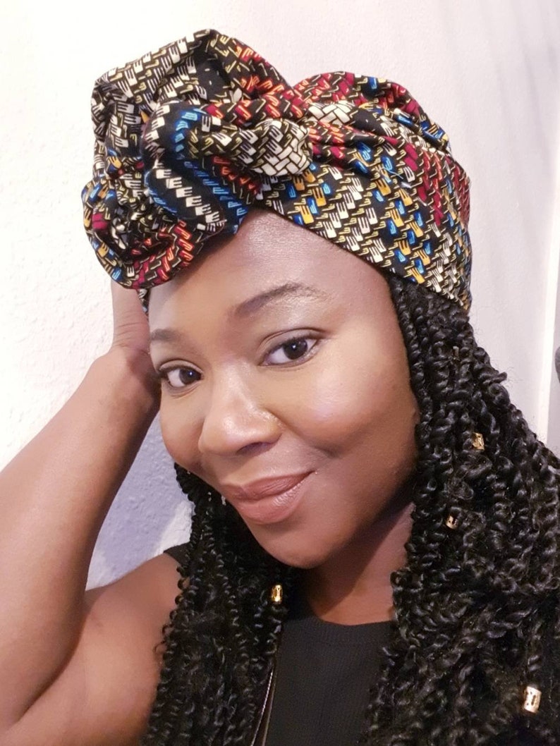 Pu&ograve; includere: Una donna con i capelli scuri e ricci indossa un turbante con motivi colorati. Il turbante &egrave; annodato sulla cima della testa e presenta un motivo geometrico con colori nero, rosso, giallo e blu.