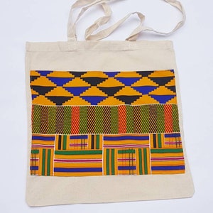 May include: A white canvas tote bag with a colorful African print design. The print features a combination of yellow, blue, green, red, and black geometric patterns.