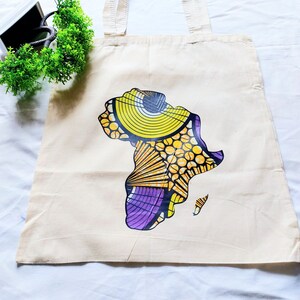 May include: A beige canvas tote bag with a colorful graphic of the continent of Africa printed on the front. The graphic features yellow, orange, purple, and black colors.