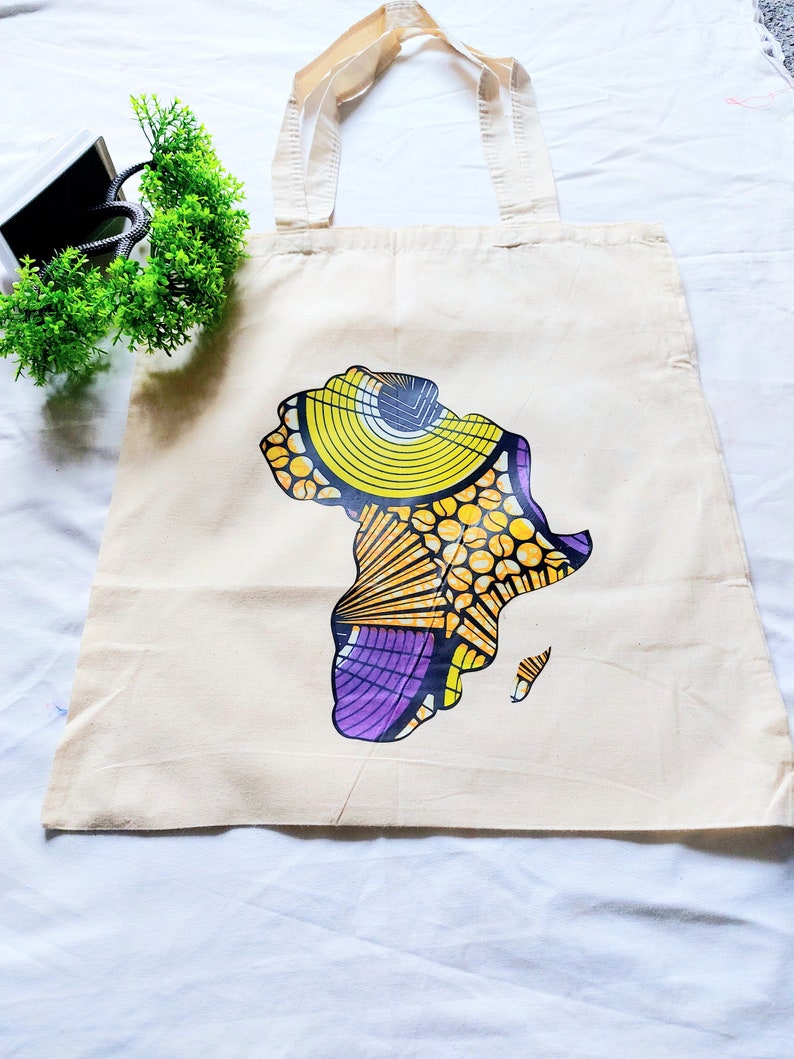 May include: A beige canvas tote bag with a colorful graphic of the continent of Africa printed on the front. The graphic features yellow, orange, purple, and black colors.
