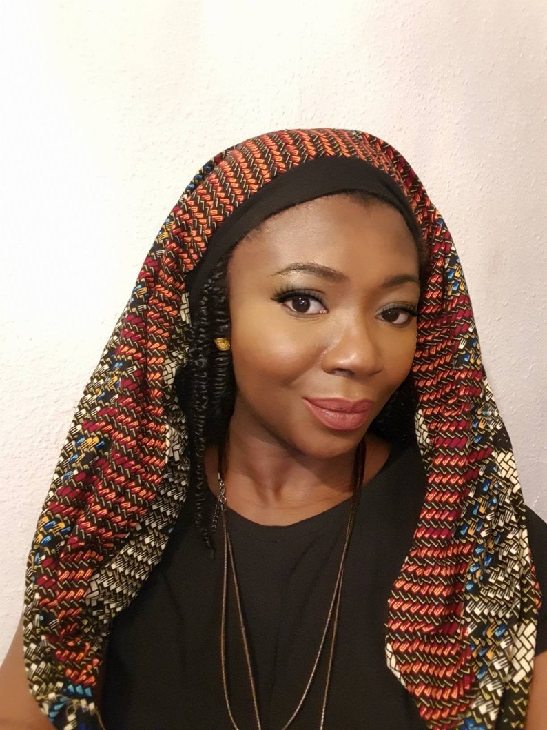 Pu&ograve; includere: Una donna indossa una camicia nera e un turbante con motivi colorati. Il turbante ha una fascia nera intorno alla fronte ed &egrave; legato dietro il collo. La donna indossa una collana.