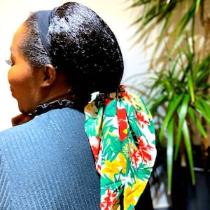 May include: A woman with black hair is wearing a black headband and a green, yellow, and red floral print scarf tied around her hair.