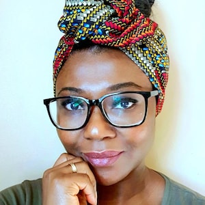 Pu&ograve; includere: Una donna con i capelli scuri raccolti in uno chignon indossa un turbante con motivi colorati con disegni geometrici rossi, gialli, blu e neri. Indossa occhiali con montatura nera e una camicia verde scuro.