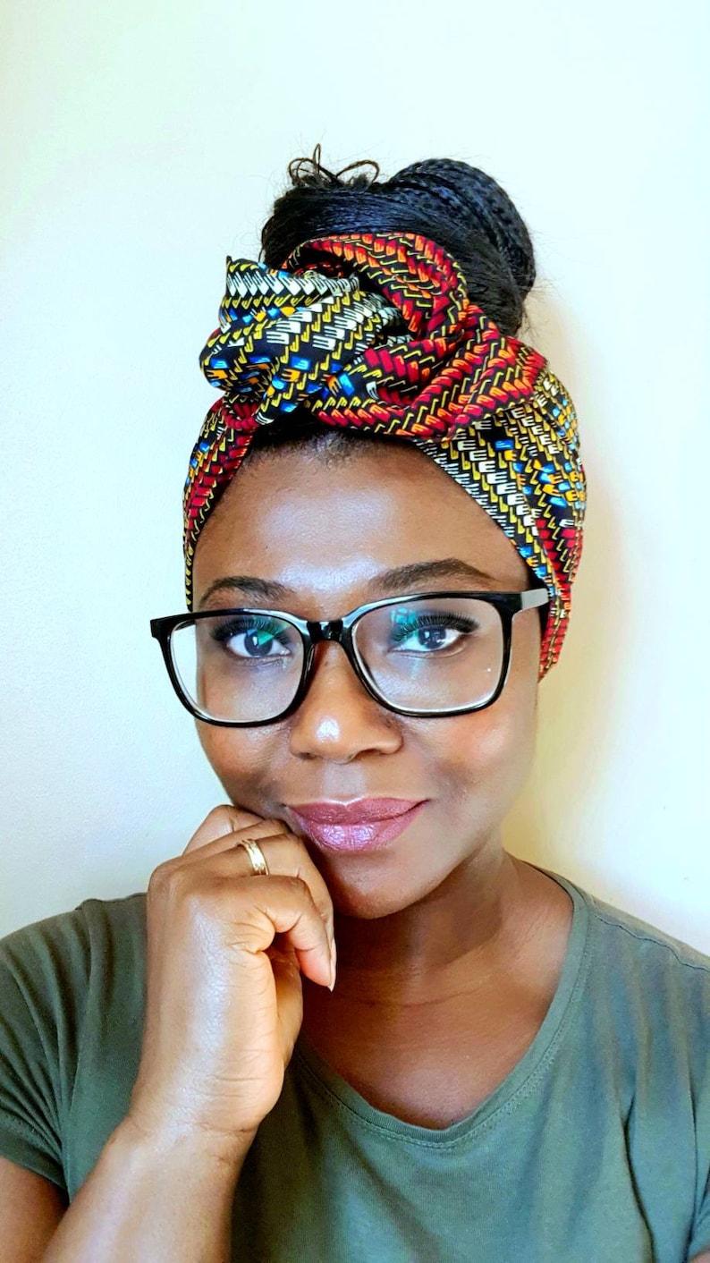 Pu&ograve; includere: Una donna con i capelli scuri raccolti in uno chignon indossa un turbante con motivi colorati con disegni geometrici rossi, gialli, blu e neri. Indossa occhiali con montatura nera e una camicia verde scuro.