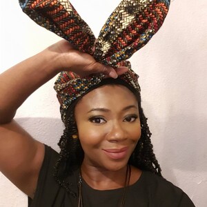 Pu&ograve; includere: Una donna indossa una camicia nera con un turbante a motivi con due estremit&agrave; lunghe e appuntite. Il turbante &egrave; legato in un nodo sulla cima della sua testa e le estremit&agrave; sono drappeggiate sulle sue spalle.