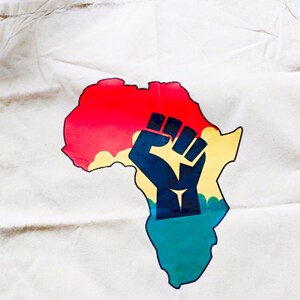 May include: A white canvas tote bag with a colorful graphic of the continent of Africa. The graphic features a raised fist in the center of the continent, with red, yellow, green, and blue colors.