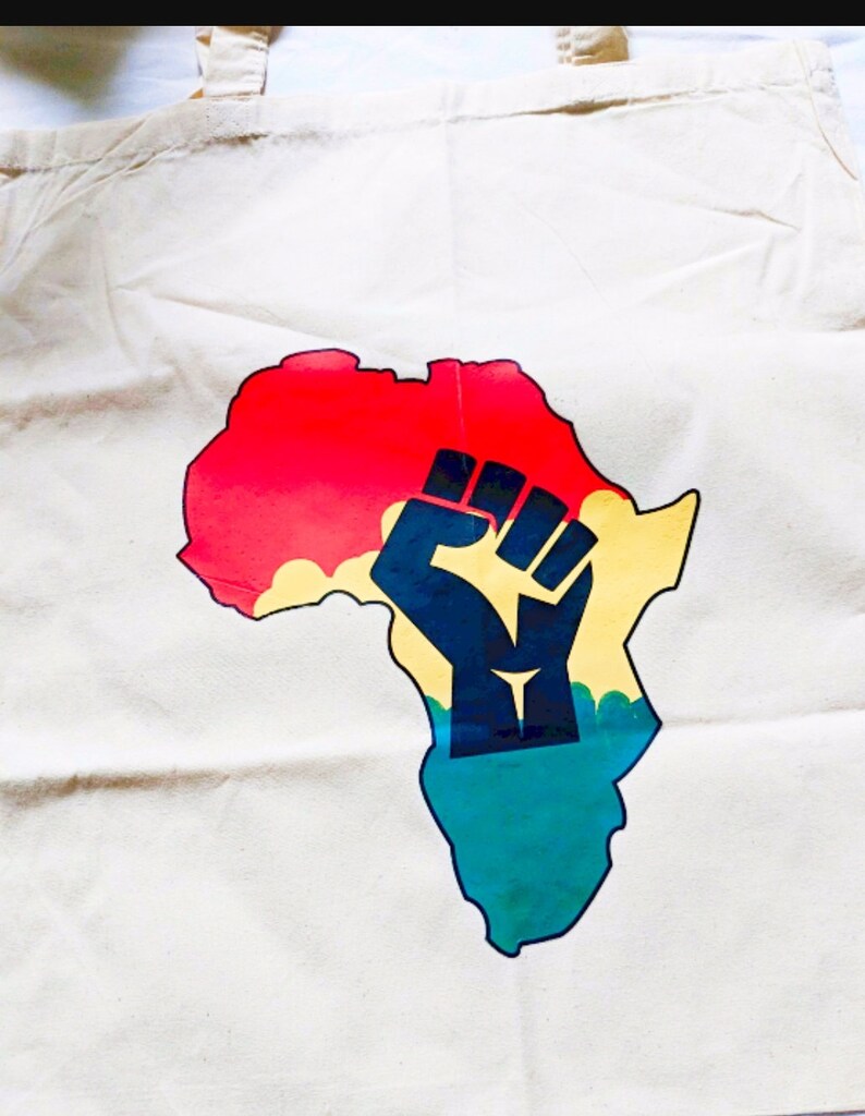 May include: A white canvas tote bag with a colorful graphic of the continent of Africa. The graphic features a raised fist in the center of the continent, with red, yellow, green, and blue colors.