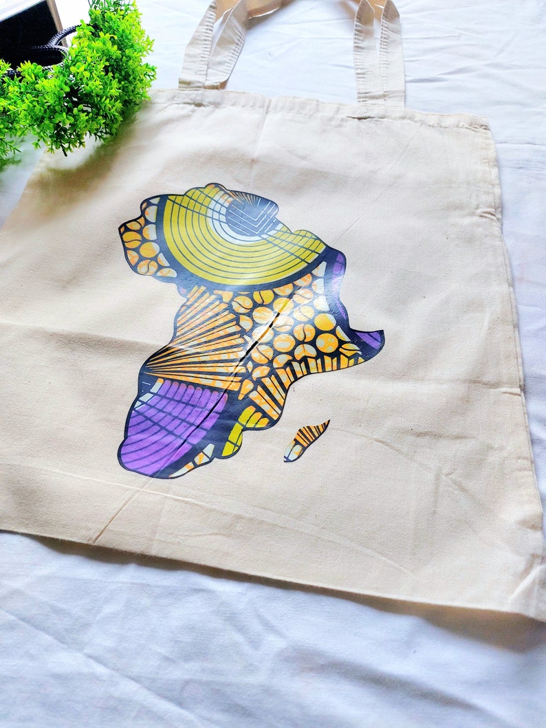 May include: A white canvas tote bag with a colorful graphic of the continent of Africa. The graphic is made up of yellow, orange, purple, and black geometric shapes.