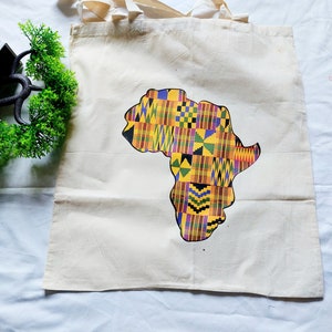 May include: A white canvas tote bag with a colorful graphic of the continent of Africa printed on it. The graphic is made up of a patchwork of yellow, red, green, blue, and black squares and rectangles.