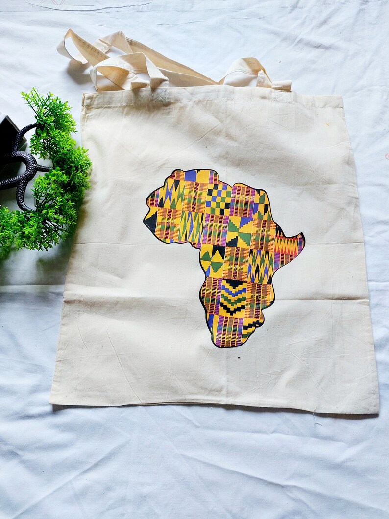 May include: A white canvas tote bag with a colorful graphic of the continent of Africa printed on it. The graphic is made up of a patchwork of yellow, red, green, blue, and black squares and rectangles.