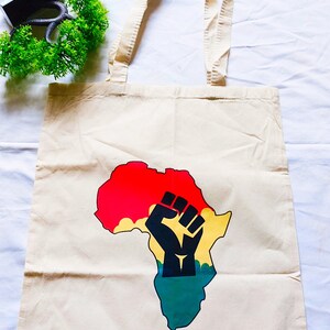 May include: A beige canvas tote bag with a black fist raised in the air over a colorful outline of the continent of Africa. The colors of the continent are red, yellow, and green.