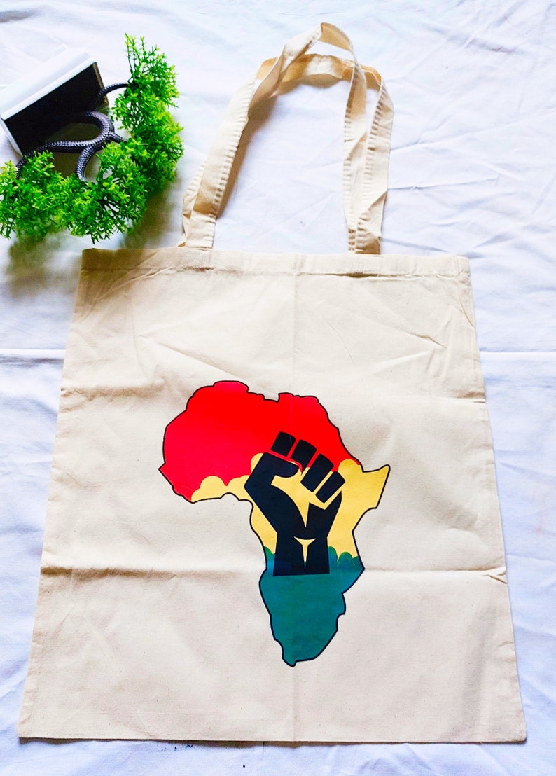 May include: A beige canvas tote bag with a black fist raised in the air over a colorful outline of the continent of Africa. The colors of the continent are red, yellow, and green.