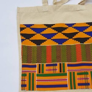 May include: A natural canvas tote bag with a large pocket on the front. The pocket is made of colorful African print fabric with geometric patterns in shades of orange, green, blue, and black.
